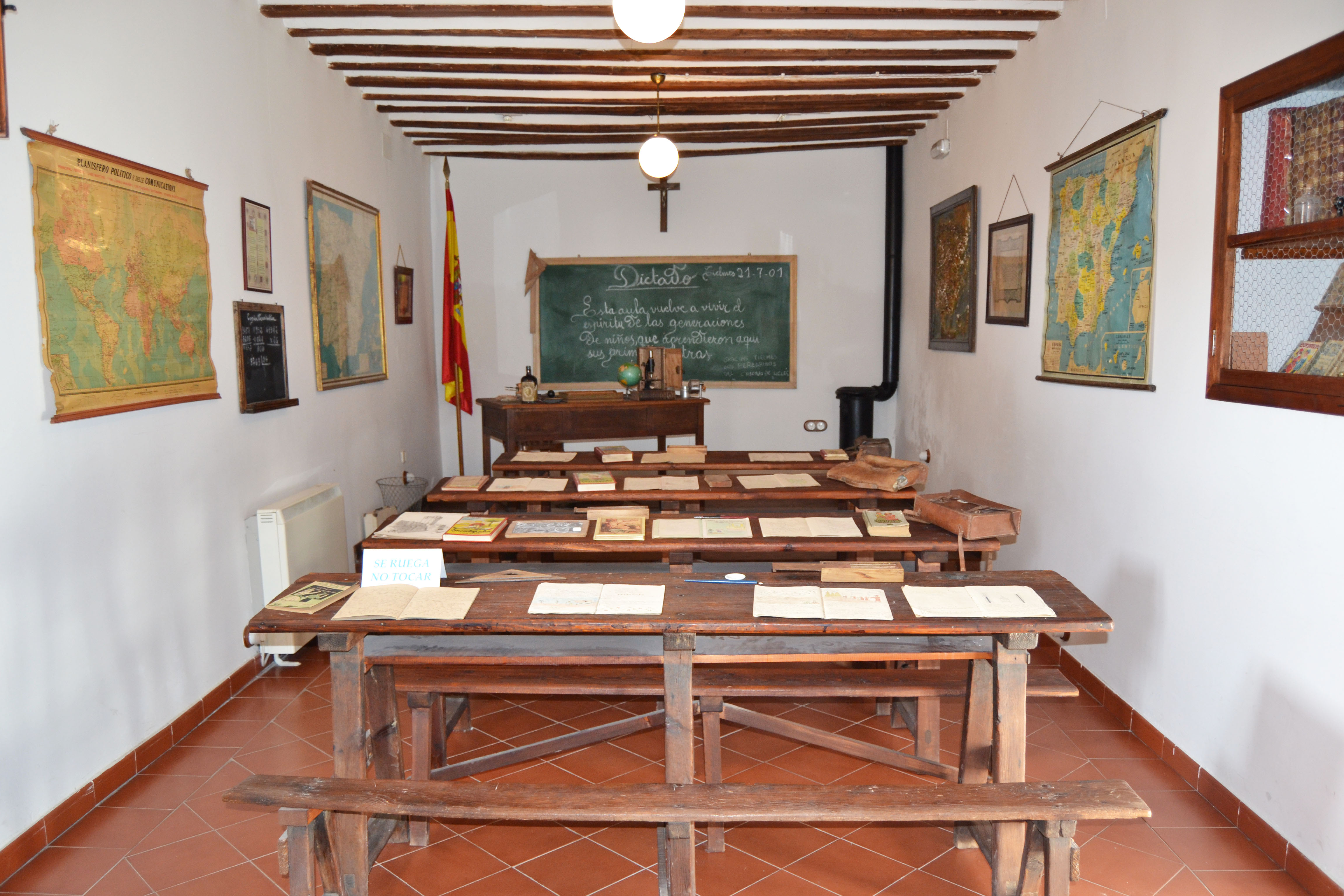 View of the interior of the house museum, showing a classroom from decades past with desks, wooden benches, pictures along the sides and a blackboard on the front wall.