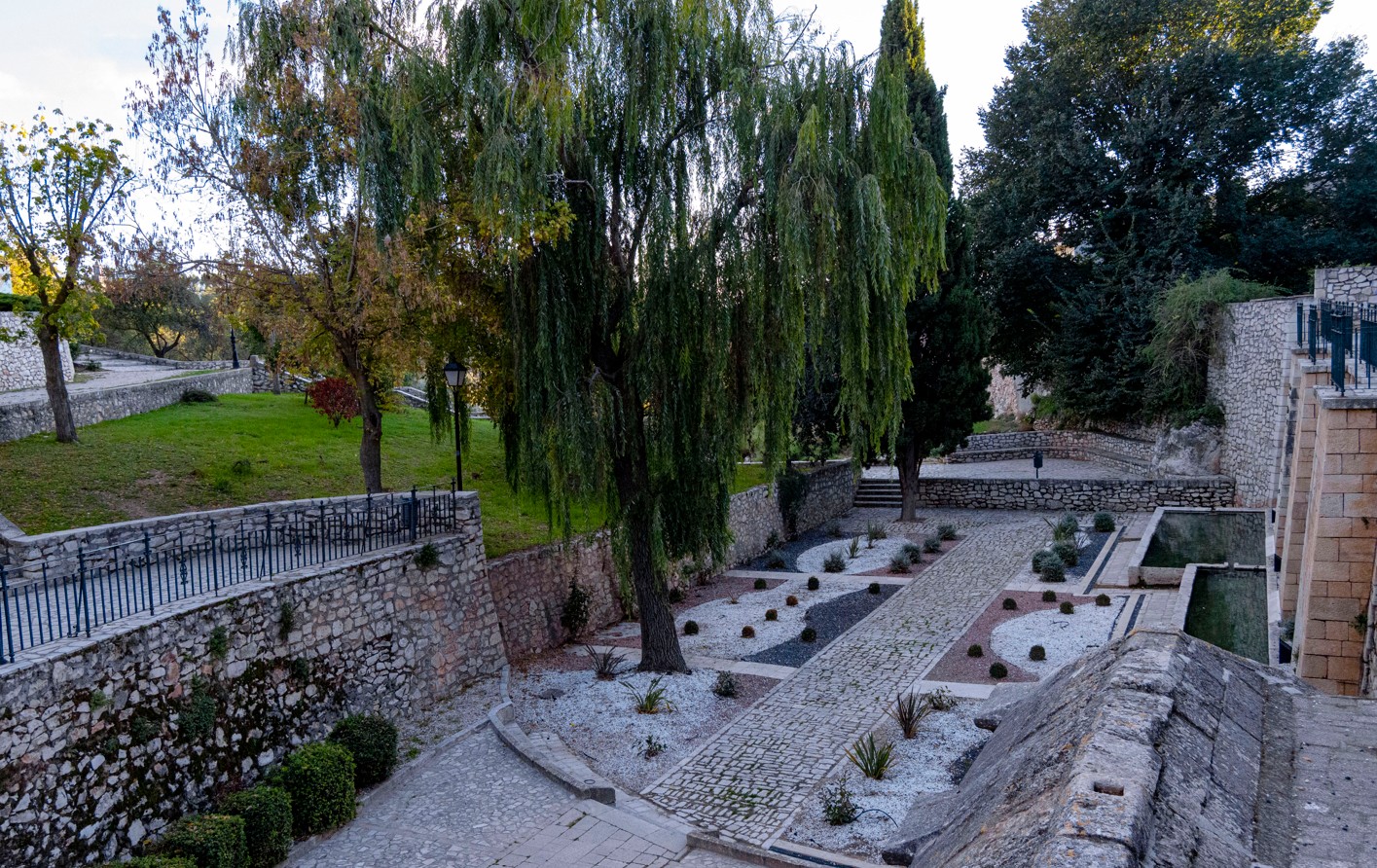 vista de los bonitos jardines, donde el protagonismo lo ganan la fuente y los pilones de piedra donde el agua llega a una robusta galería de cantería para verter por un solo caño en una amplia caja cubierta por un entramado de losas apoyadas sobre arcos románicos. De esta primera galería pasa el agua a otro pilón descubierto y luego a otro.