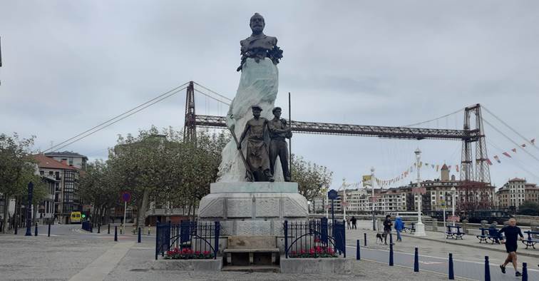 Escultura de unos 8 metros de altura coronada por un busto sobre un pedestal al frente del cual hay dos hombres