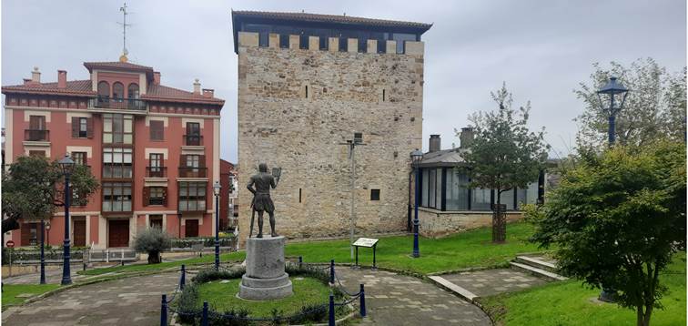 Gizon irakurtzen ari den eskultura ikus daitekeen plaza xarmangarri baten atzean, harlanduzko dorretxe karratu bat dago.