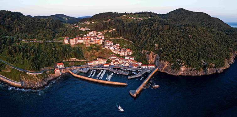 Foto de montañas verdes que terminan en acantilados en un mar de intenso azul y en el centro un puerto al abrigo de dos diques. En el acantilado que baja al puerto se ven casas que trepan la ladera formando un pintoresco pueblecito.
