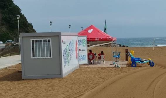 Foto en la que se ve una caseta de asistencia al baño, un toldo para sombra, con sillas y dos personas, y una silla anfibio en la arena.