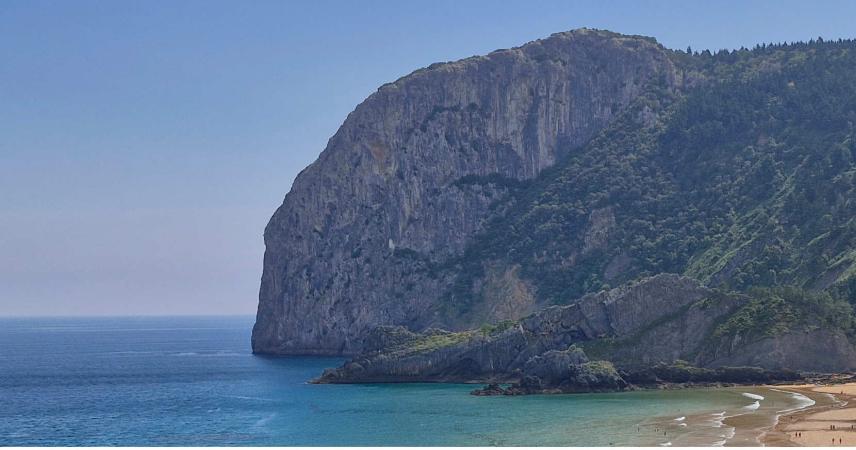 Photo d'un énorme rocher de pierre grise, avec des arbres vert foncé à sa droite, et à sa gauche et en dessous une mer bleue qui atteint une plage de sable doré à droite