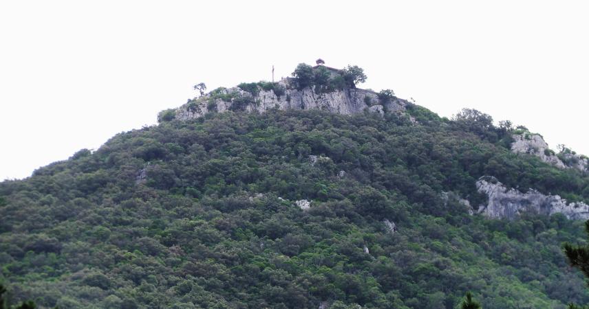 Photographie d'une montagne verte et boisée avec un sommet rocheux, où l'on peut voir une petite chapelle.