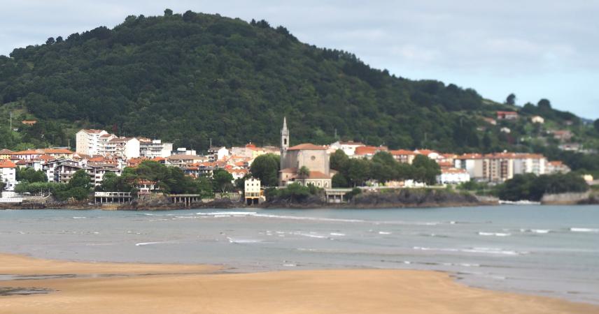 Foto de un pueblo a la orilla del mar, con una colina verde al fondo y arbolado en primera línea. Casas de varias alturas con tejados rojos. En el centro destaca una iglesia.
