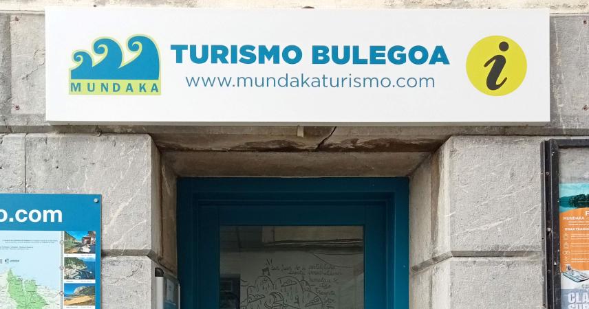 Foto de la puerta de entrada de la oficina de turismo, está enmarcada en azul sobre fachada de piedra gris. No hay escalones.